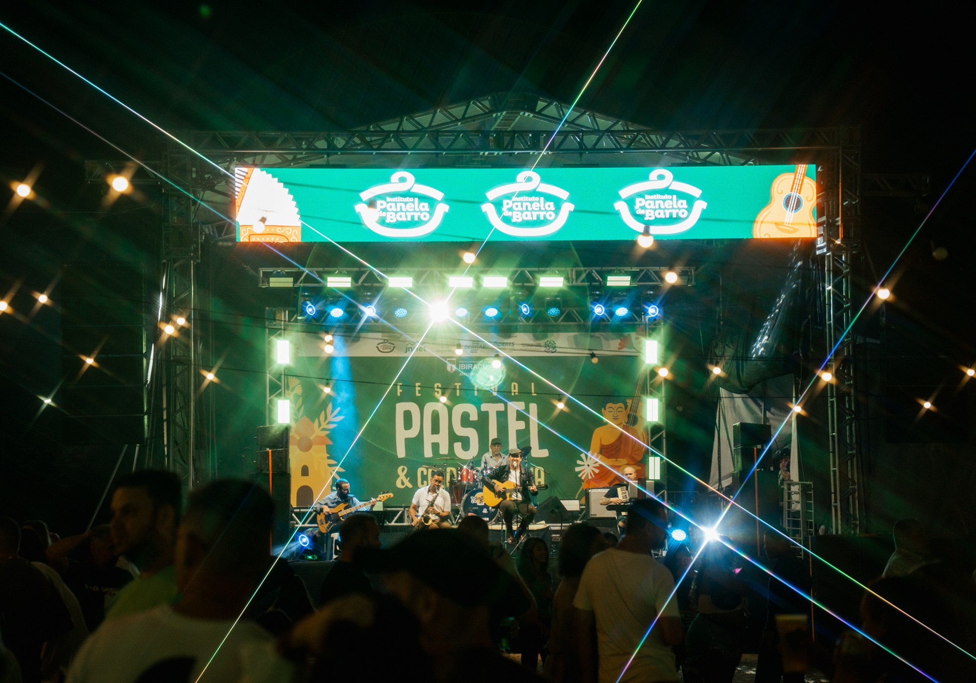 Palco iluminado do Festival do Pastel e Caldo de Cana, com músicos e público na edição anterior.