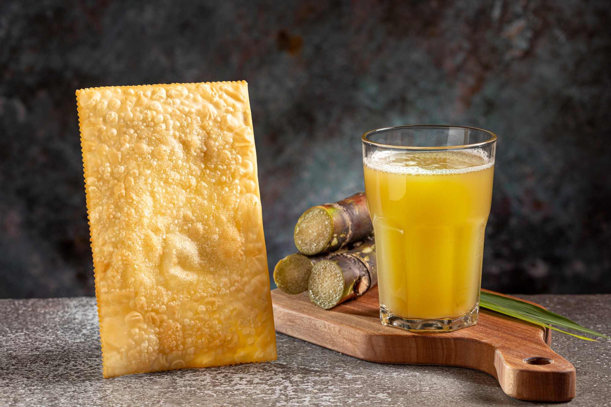 Pastel dourado ao lado de um copo de caldo de cana e pedaços de cana-de-açúcar.