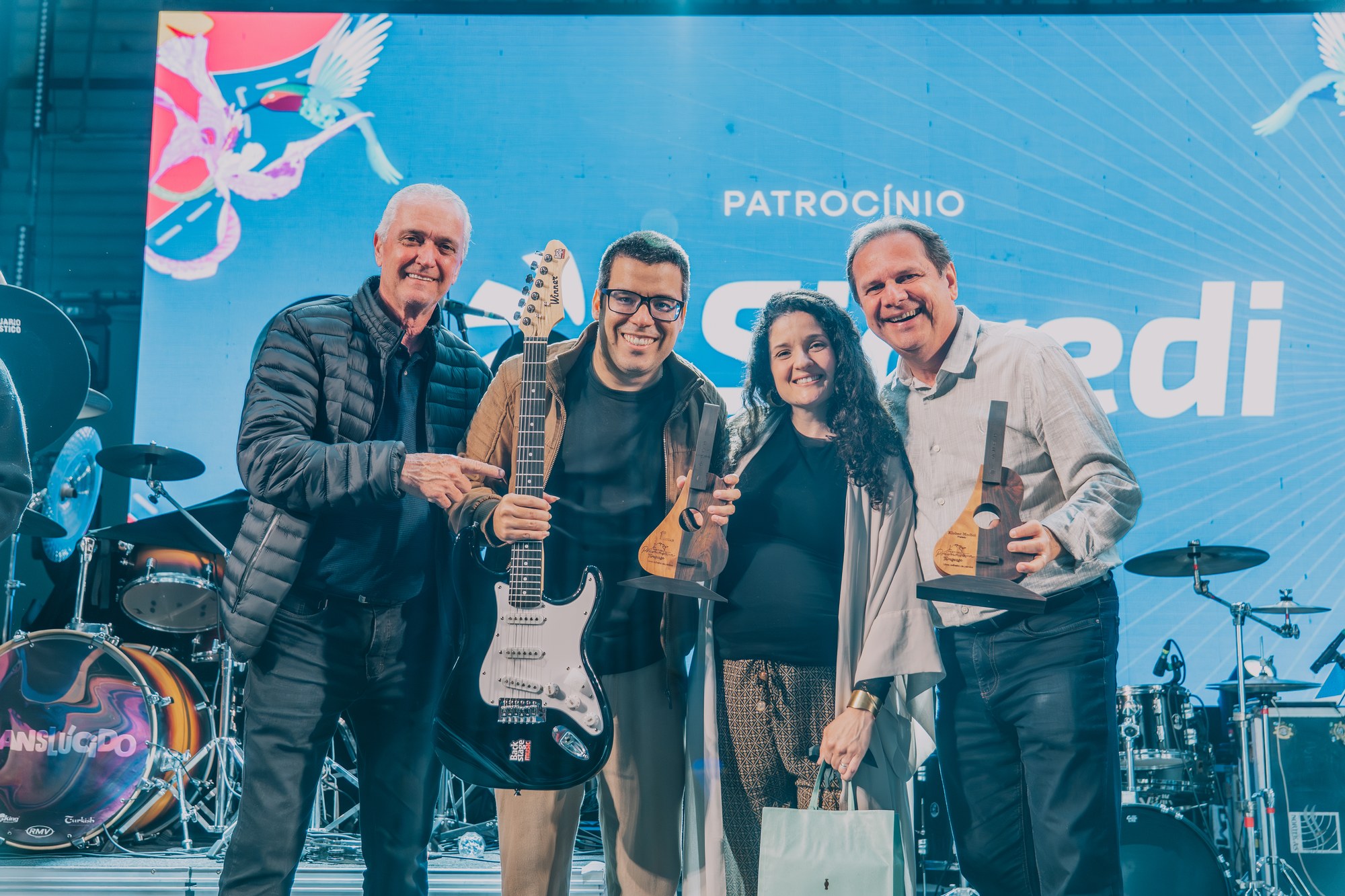 Participantes reunidos com troféus e guitarra no palco da final do concurso de música.